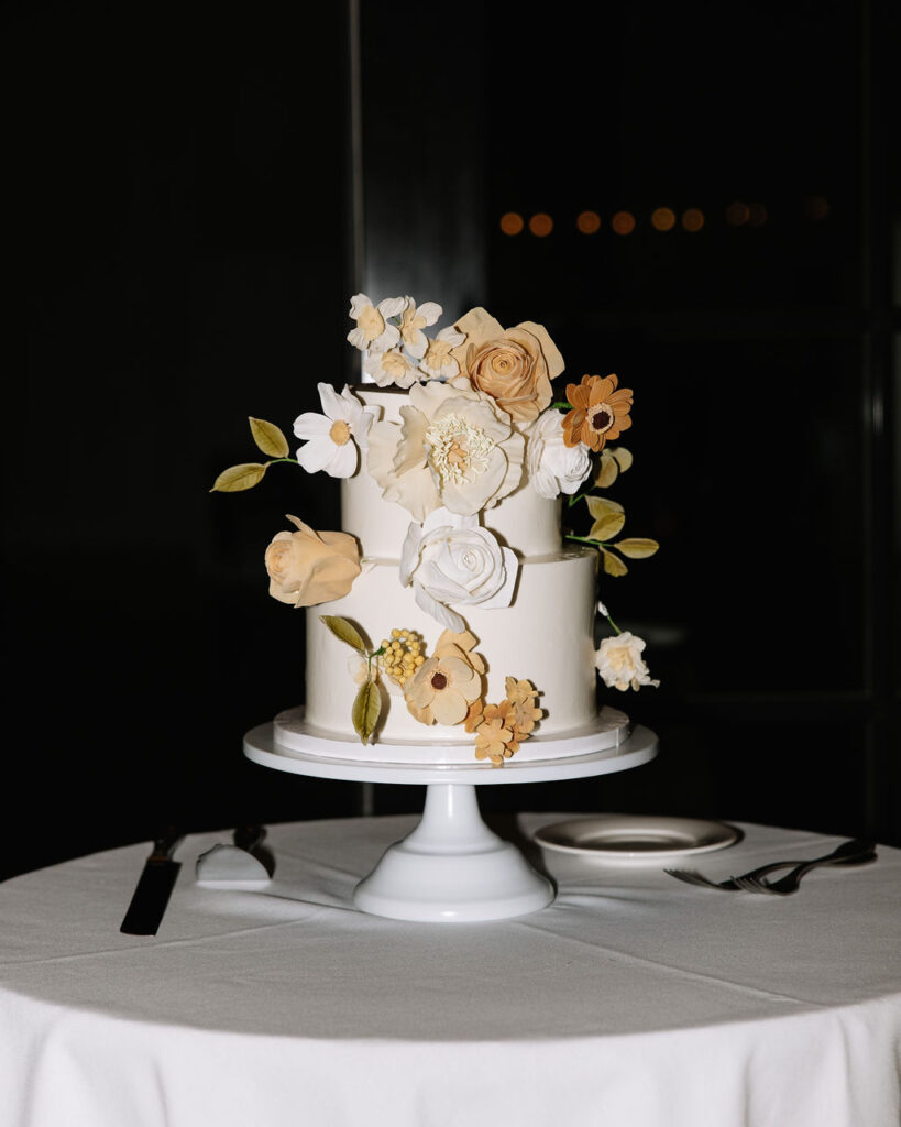 A two-tiered white cake decorated with beige, yellow, and white sugar flowers sits on a white cake stand on a round table with utensils and a plate.