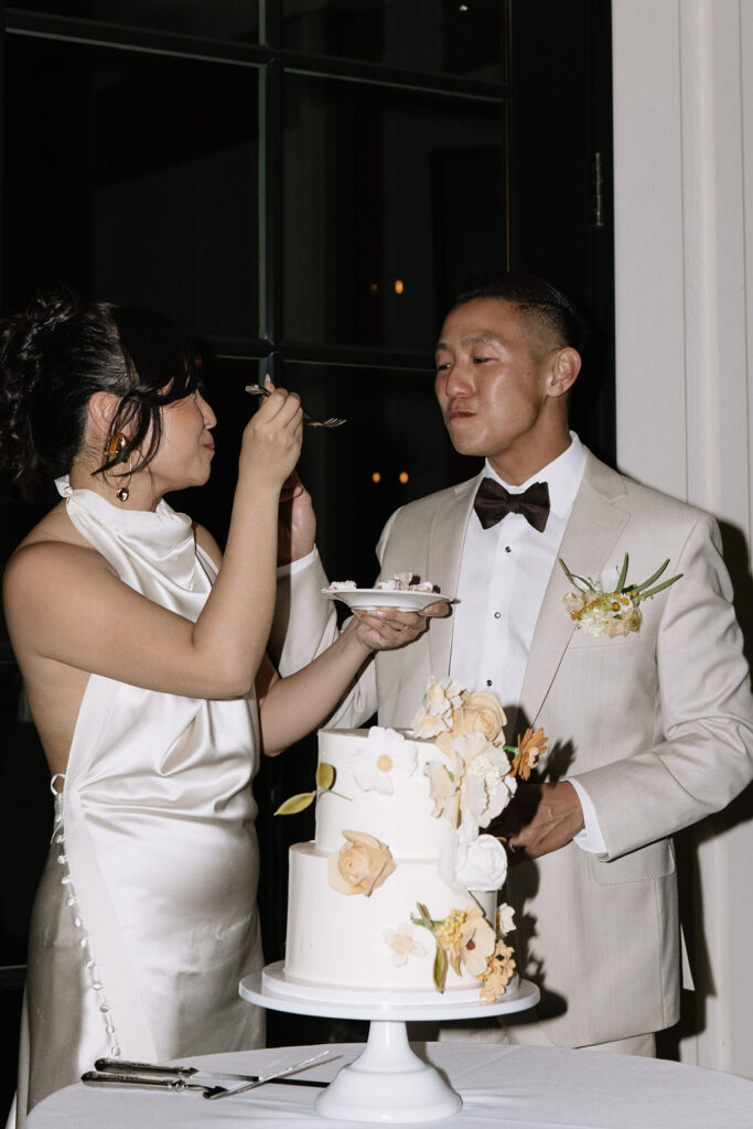 A bride in a white satin dress feeds cake to a groom in a light suit with a bow tie, standing beside a decorated tiered wedding cake.