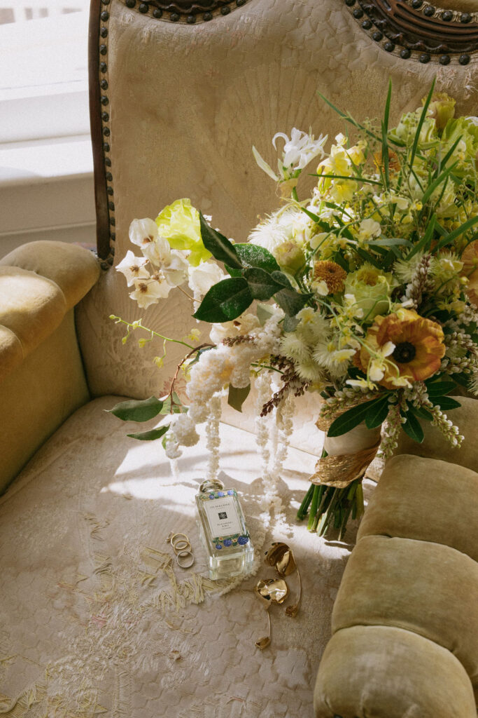 A beige upholstered chair with a bouquet of flowers, a bottle of Jo Malone perfume, gold rings, and a gold bracelet resting on the seat.