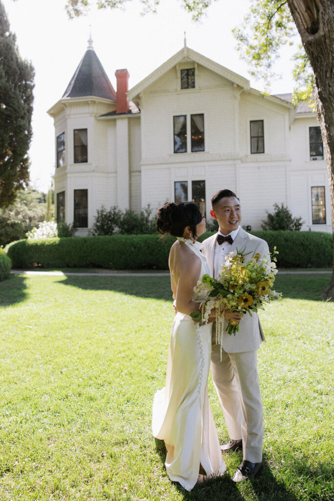 A bride and groom stand together on a lawn in front of a large white house; the bride holds a bouquet and wears a white dress, the groom wears a light-colored suit and bow tie.