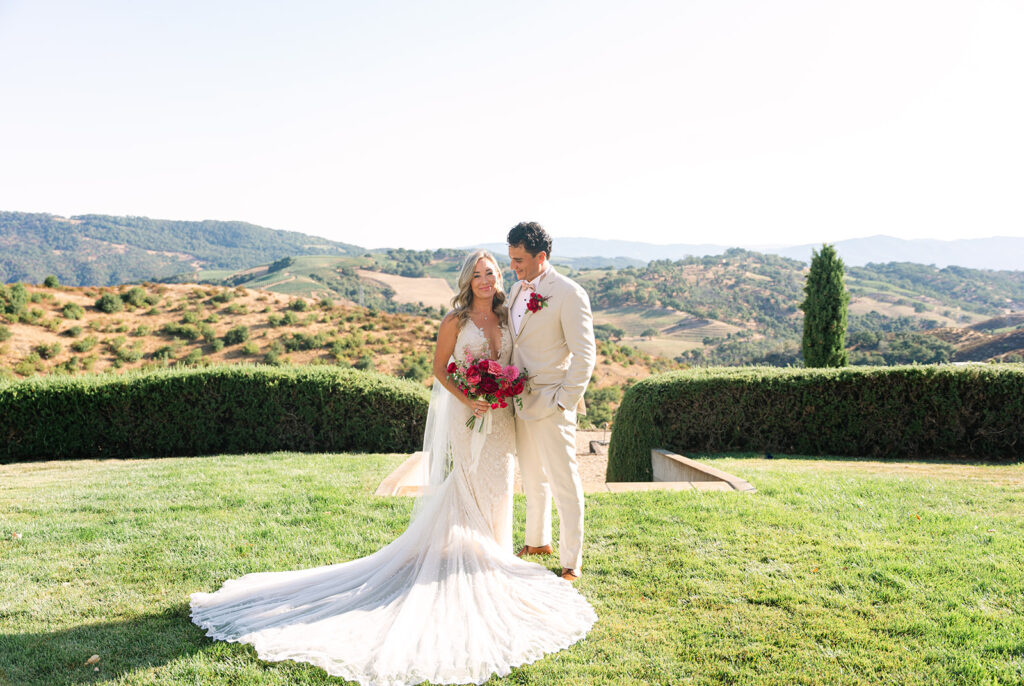 A bride and groom in formal attire stand together on a lawn, with green hills and clear skies in the background. The bride holds a bouquet of red flowers.