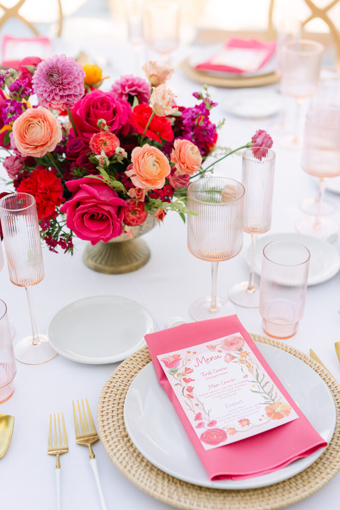 A table setting with a floral centerpiece of pink and orange flowers, pink glassware, gold cutlery, and a menu placed on a white plate with a woven charger.