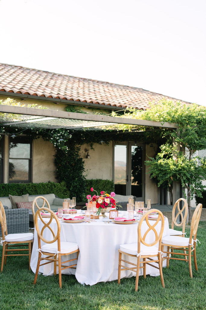 A round outdoor dining table with a white tablecloth, wooden chairs, and a floral centerpiece is set on a lawn near a patio and a vine-covered house.