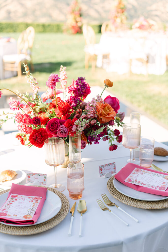A round table set for two with gold utensils, pink napkins, menus, place cards, and a vibrant floral centerpiece in red, pink, and orange hues.