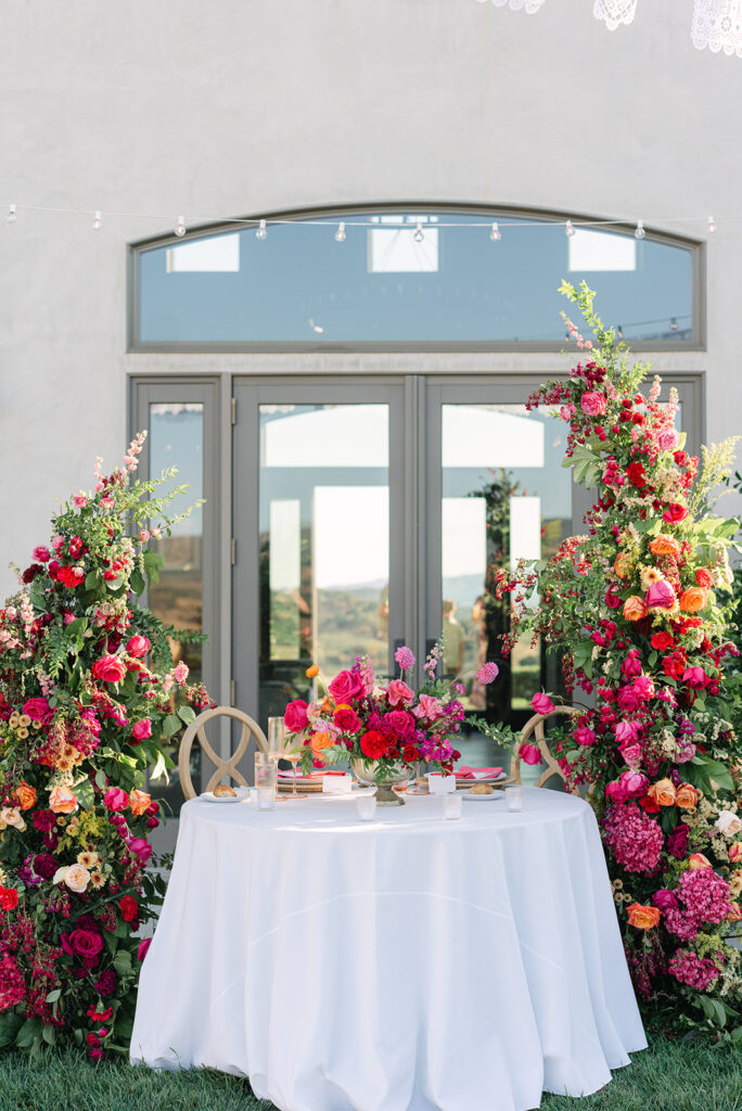 A round table with a white tablecloth is set for two in front of glass doors, surrounded by vibrant, colorful floral arrangements.