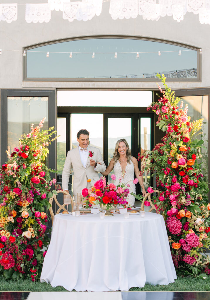 A bride and groom stand smiling behind a table with a white tablecloth, surrounded by vibrant red, pink, and orange floral arrangements in an outdoor setting.