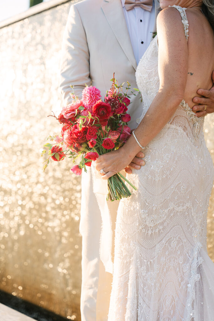 A bride in a lace dress holds a bouquet of red and pink flowers, standing next to a groom in a light suit in front of a golden textured background.