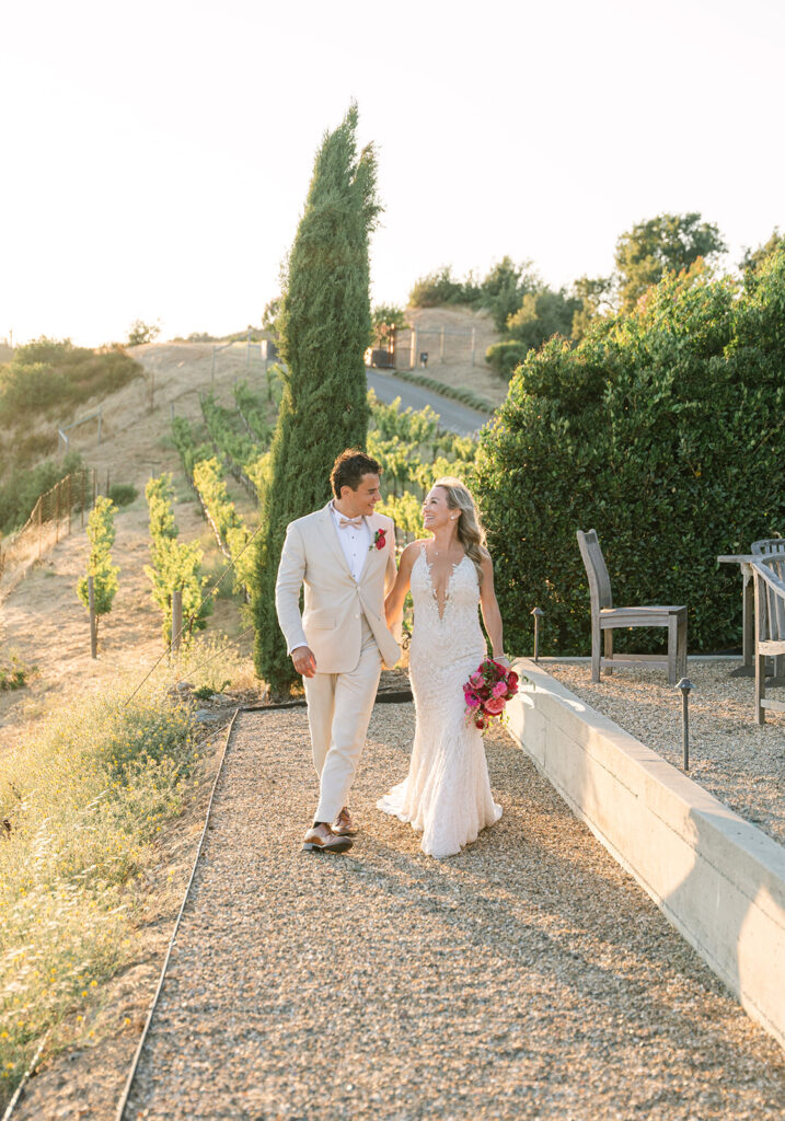 A bride and groom walk outside along a gravel path at a vineyard, holding hands and smiling at each other in the sunlight.