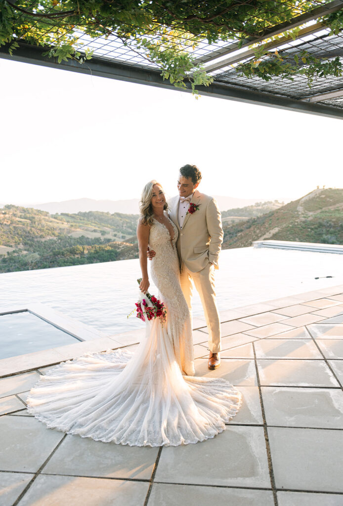 A bride and groom stand together by an outdoor pool, with hills in the background. The bride wears a long, fitted gown and holds a bouquet; the groom wears a light suit with a red boutonniere.