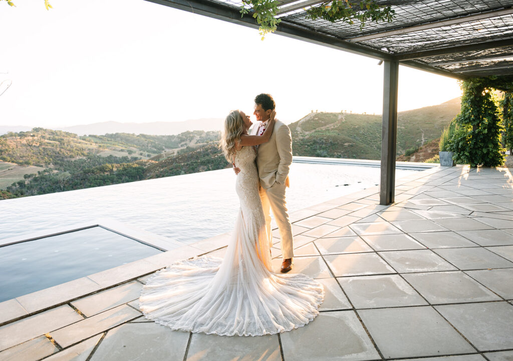 A bride and groom stand embracing by an infinity pool on a tiled terrace overlooking hilly countryside at sunset.