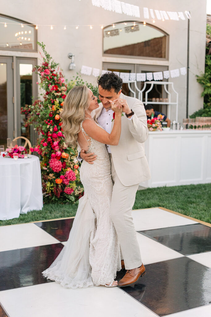 A bride and groom dance together on a black and white checkered floor at an outdoor wedding reception, surrounded by colorful floral decorations.