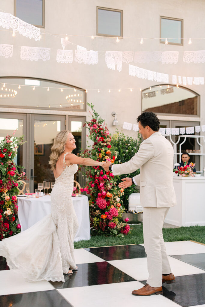 A bride and groom dance together on a black and white checkered floor at an outdoor wedding, with colorful flowers and string lights in the background.