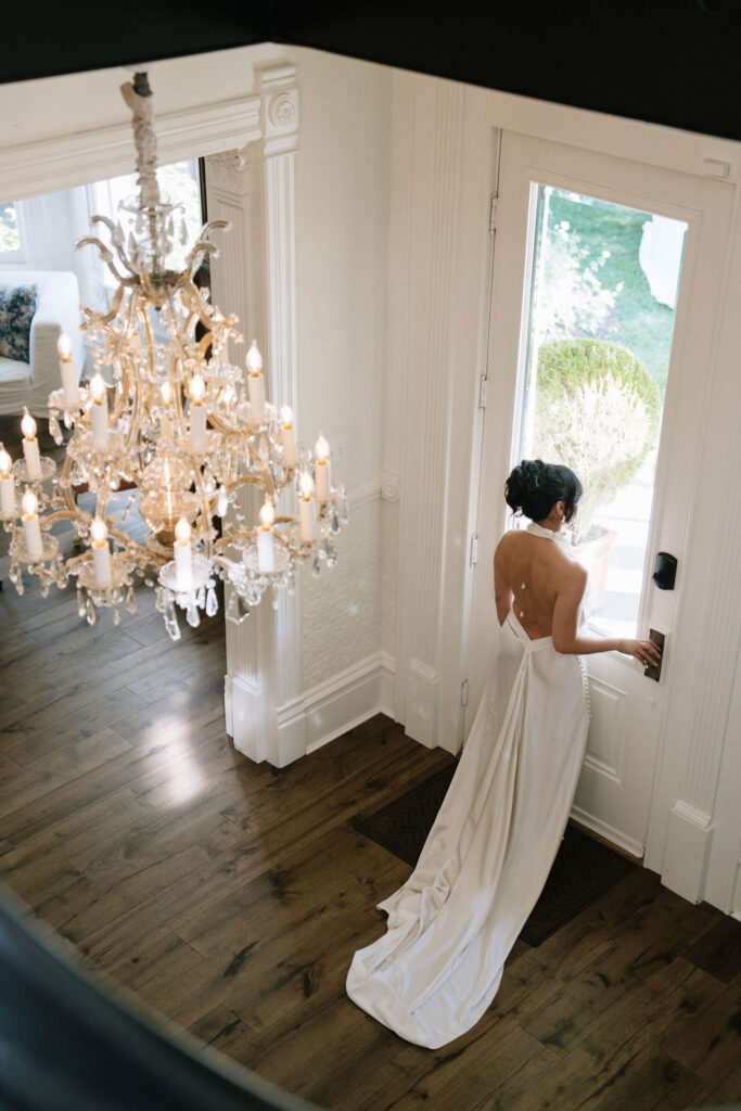 A woman in a white backless gown stands at a door in a bright, elegant room with a large chandelier and wooden floors.