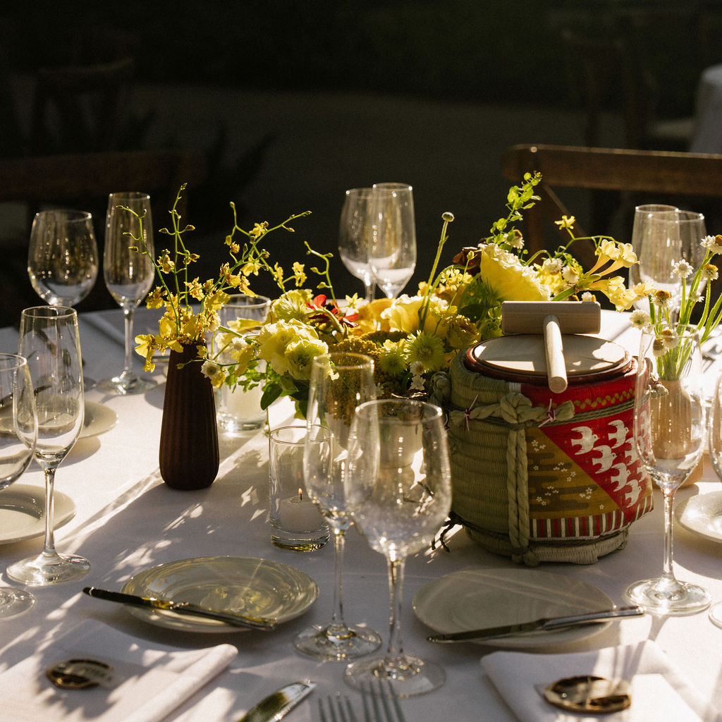 A sunlit table set for dining with empty wine glasses, plates, napkins, yellow flowers, and a decorative cheese plate on a drum centerpiece.