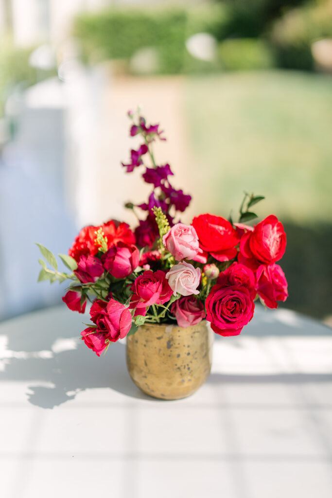 A gold vase filled with red and pink roses and other flowers sits on a white table outdoors, with greenery blurred in the background.