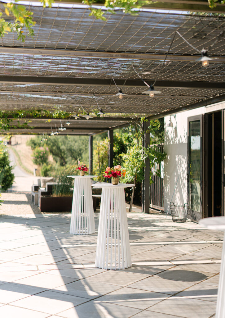 An outdoor patio with tall white cocktail tables, each topped with a small floral arrangement, under a pergola with string lights.