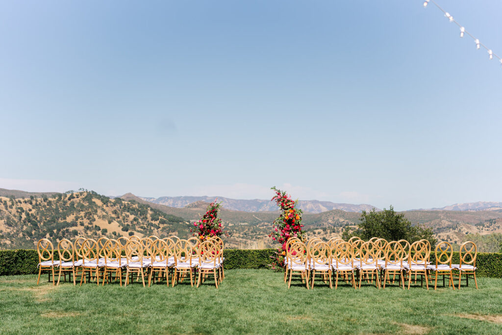 Rows of wooden chairs arranged outdoors on a lawn, facing a floral arch, with mountains and a clear sky in the background.