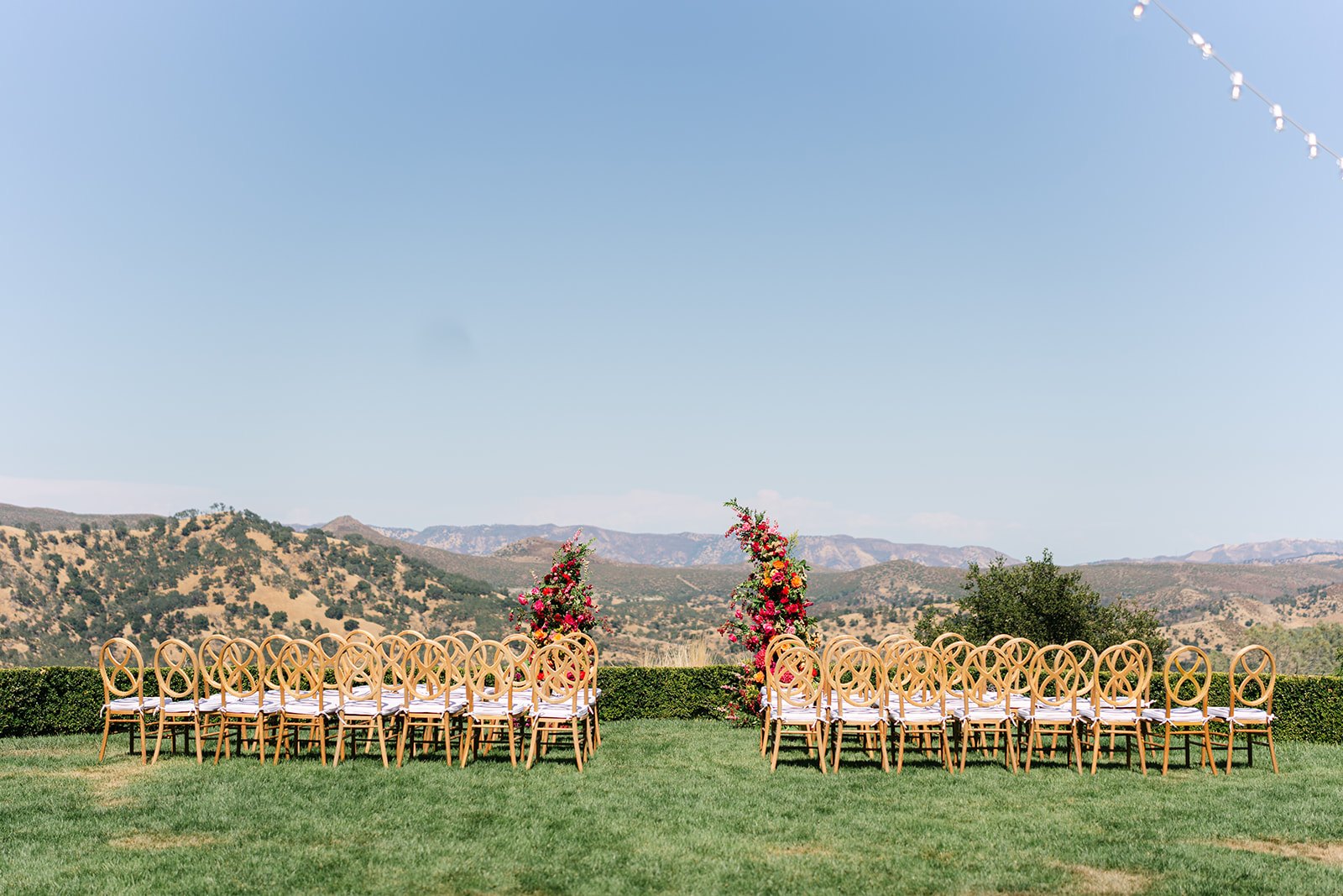 Rows of wooden chairs arranged outdoors on a lawn, facing a floral arch, with mountains and a clear sky in the background.