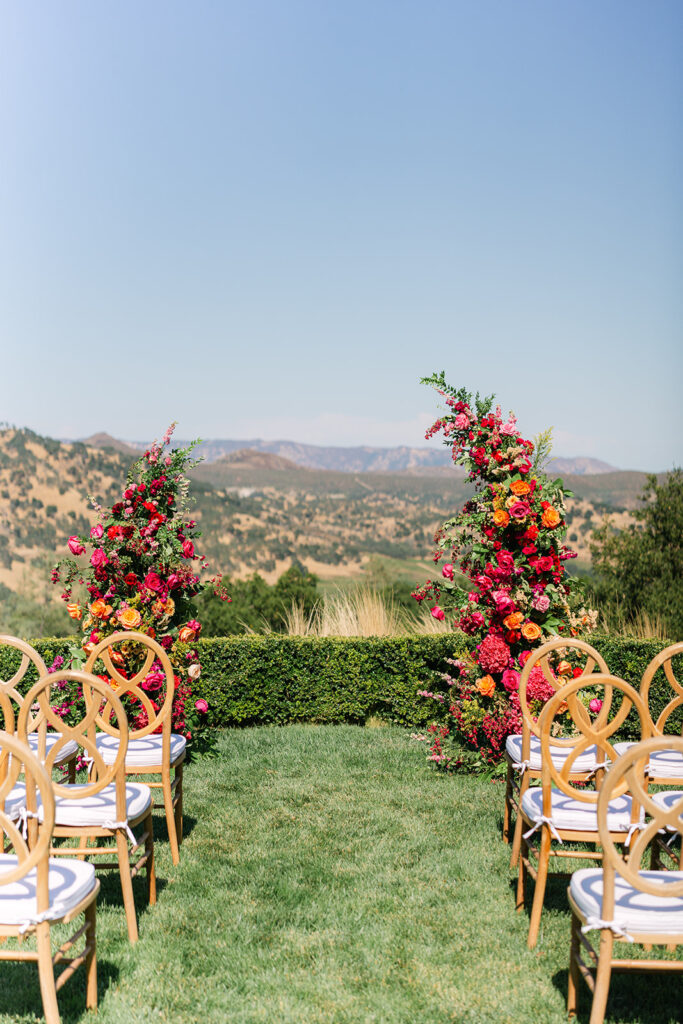 Outdoor wedding ceremony setup with rows of wooden chairs facing two colorful floral arches, overlooking a scenic mountain landscape under a clear blue sky.