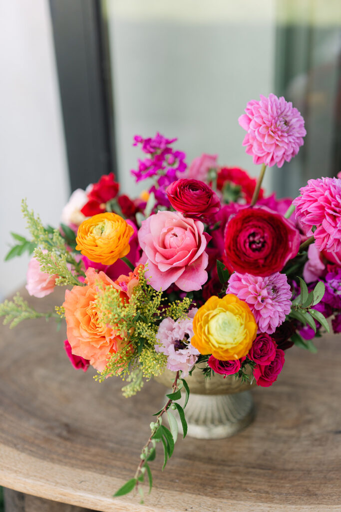 A floral arrangement in a silver vase featuring pink roses, yellow, orange, and red ranunculus, dahlias, and green foliage on a wooden table.