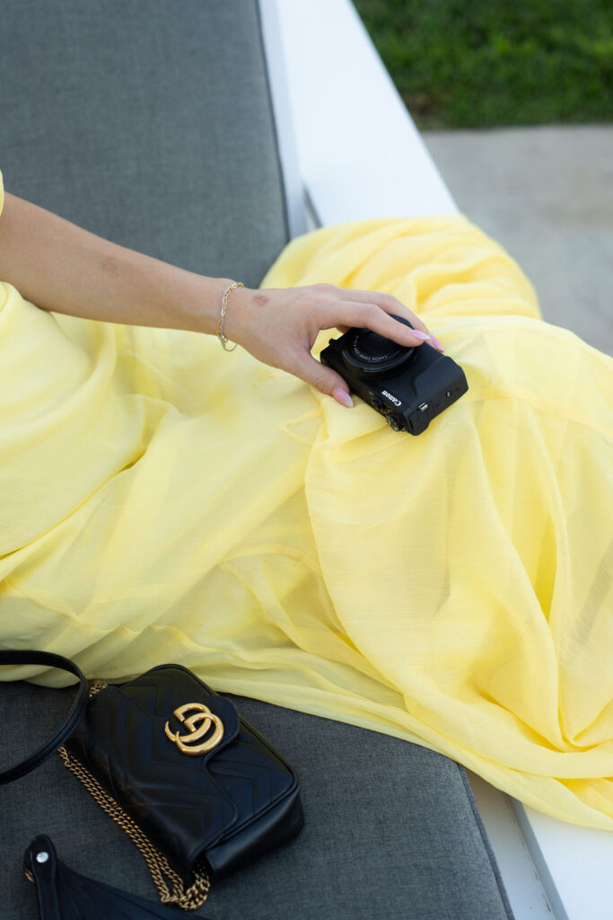 A person in a yellow dress holds a black camera while sitting on a gray surface next to a black purse with a gold chain.
