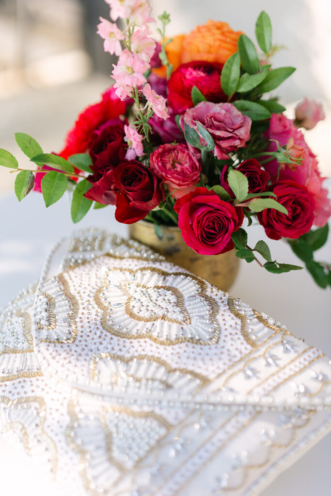 A beaded white and gold clutch rests on a table next to a vase of assorted vibrant flowers including roses and greenery.