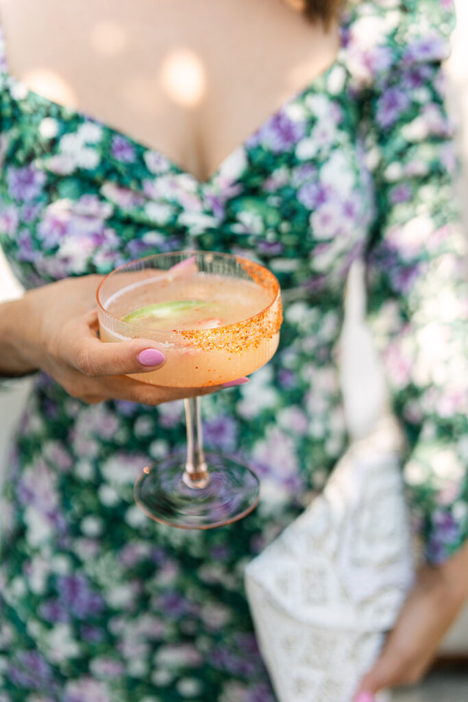 A person in a green and purple floral dress holds a cocktail glass with an orange rim and a light drink, with a white textured clutch in their other hand.