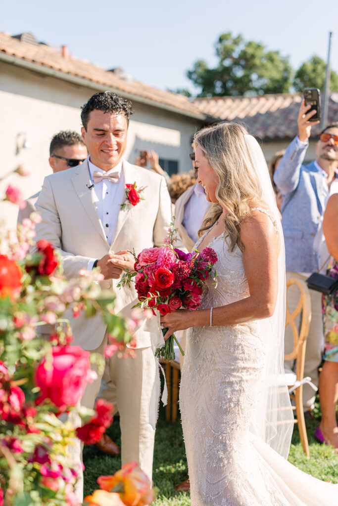 A bride in a lace dress holds a bright bouquet and stands beside a groom in a beige suit during an outdoor wedding ceremony.