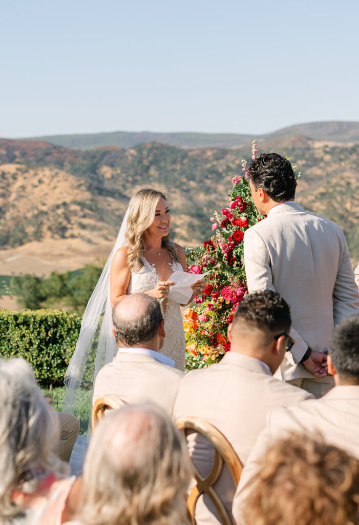 A bride and groom stand facing each other outdoors during their wedding ceremony, with guests seated and a colorful floral arrangement behind them.