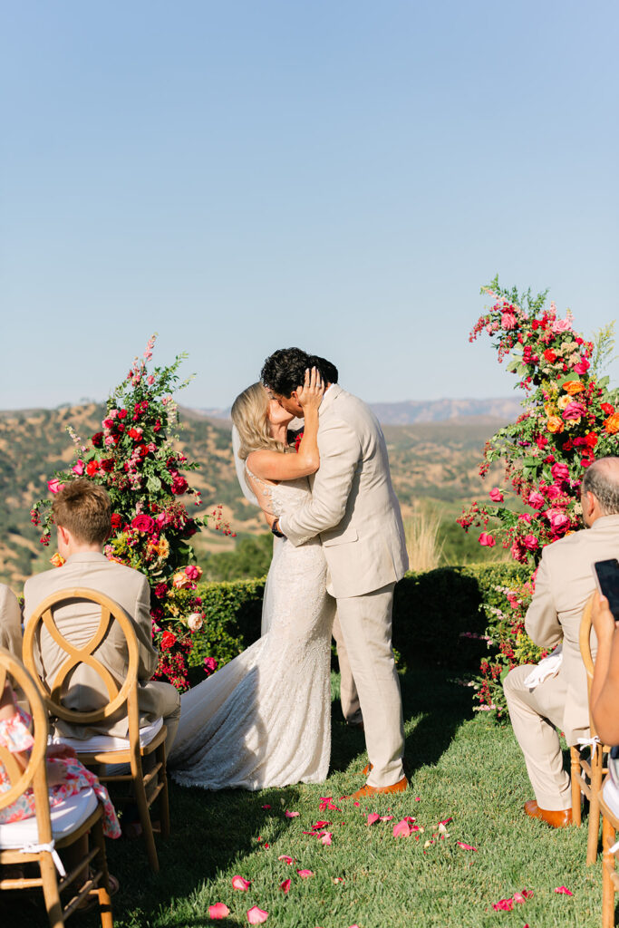 A bride and groom kiss at an outdoor wedding ceremony, surrounded by colorful floral arrangements and seated guests, with mountains in the background.