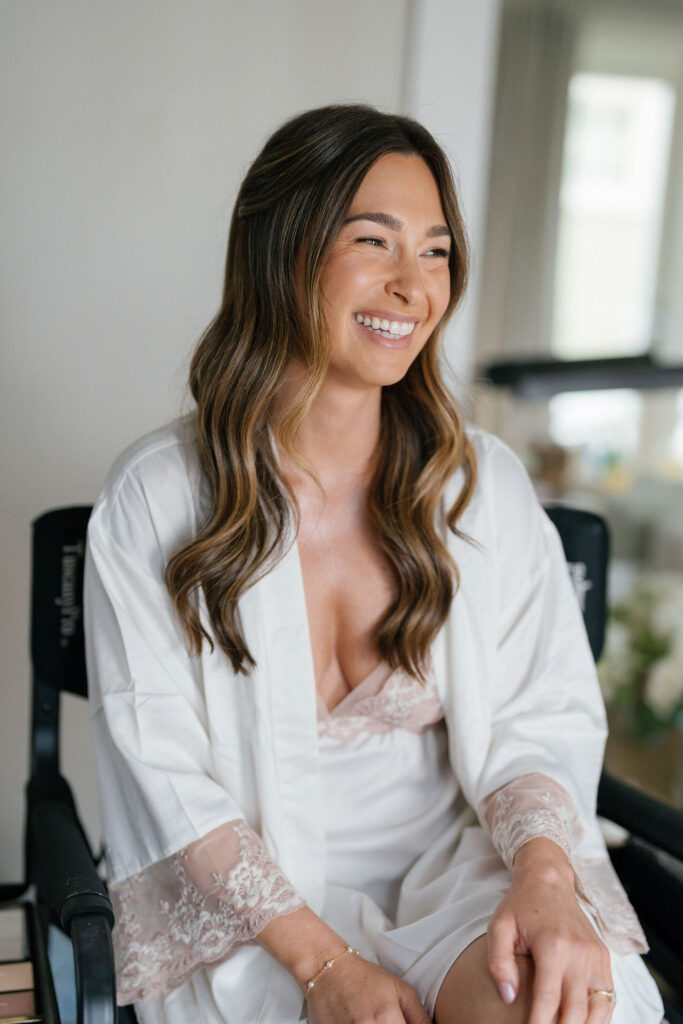 A woman with long brown wavy hair, wearing a white robe with lace cuffs, sits and smiles indoors.