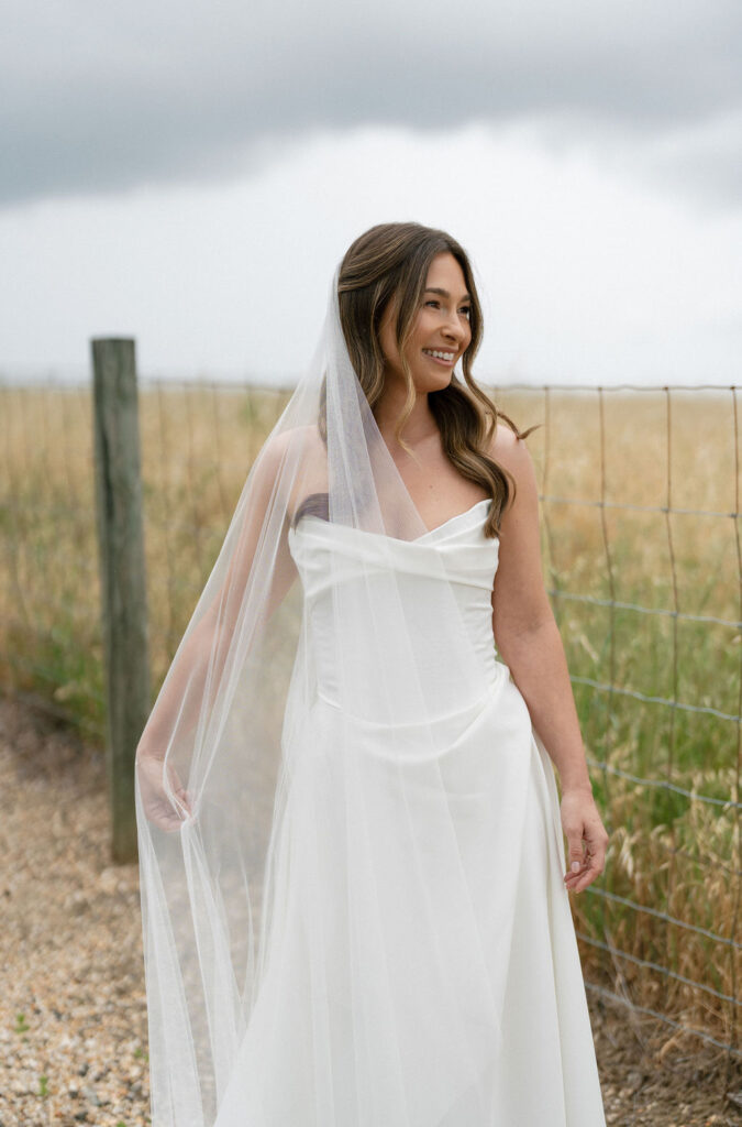 A woman in a white wedding dress and veil stands outdoors by a wire fence, with grass and cloudy sky in the background.