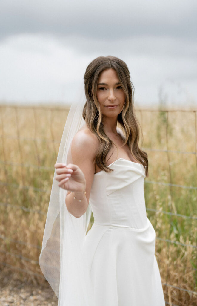 A woman in a white strapless wedding dress and veil stands outdoors near a wire fence, with tall grass and a cloudy sky in the background.
