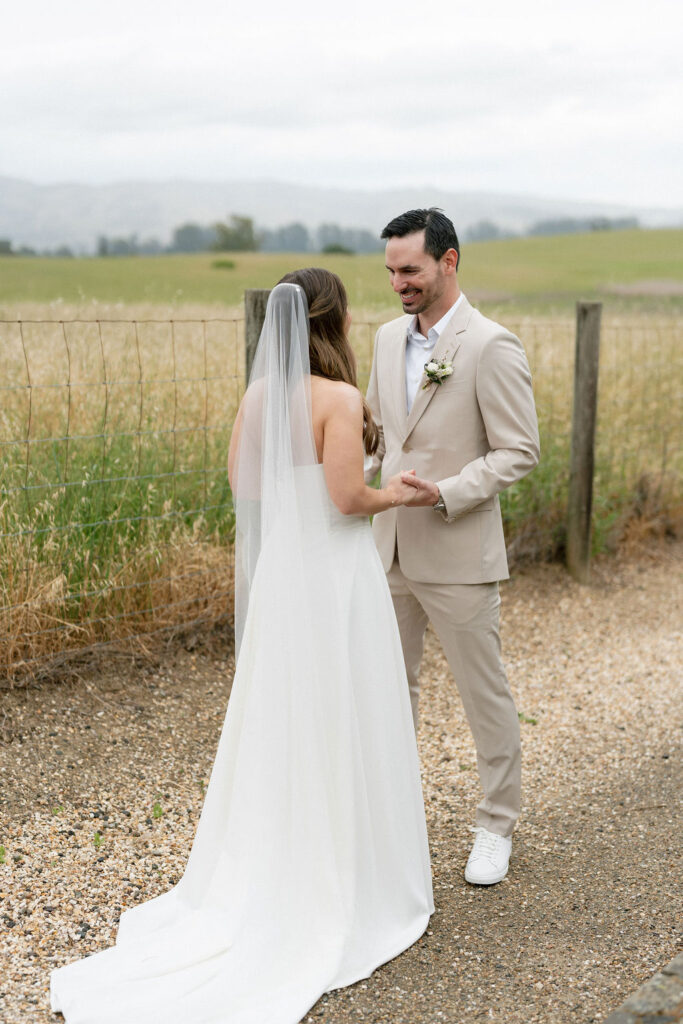 A bride and groom stand outdoors on a gravel path, holding hands and facing each other, with a grassy field and mountains in the background.