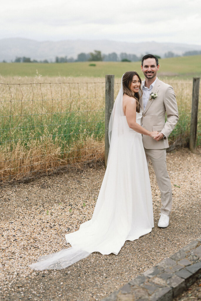 A bride and groom stand side by side outdoors on a gravel path, holding hands and smiling, with fields and mountains in the background.
