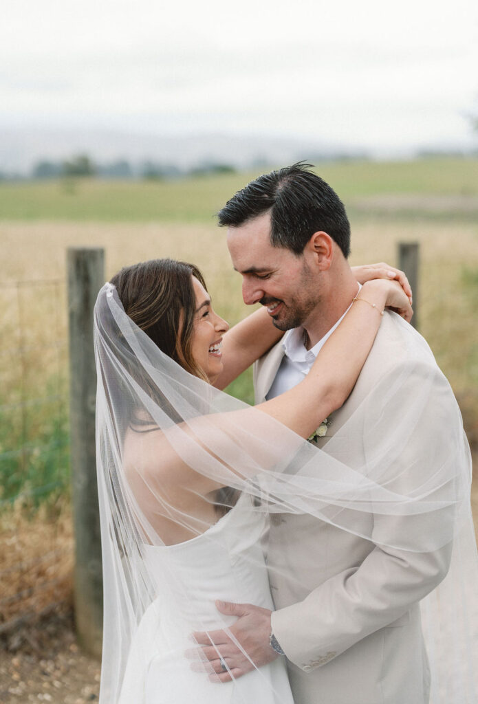 A bride and groom embrace and smile at each other outdoors, with the bride's veil draped around both of them.