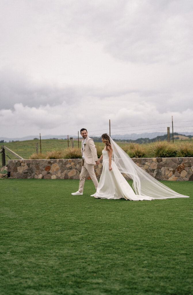 A bride and groom walk hand in hand across a grassy lawn, with the bride wearing a long veil and the groom in a light-colored suit, against a cloudy sky and stone wall backdrop.
