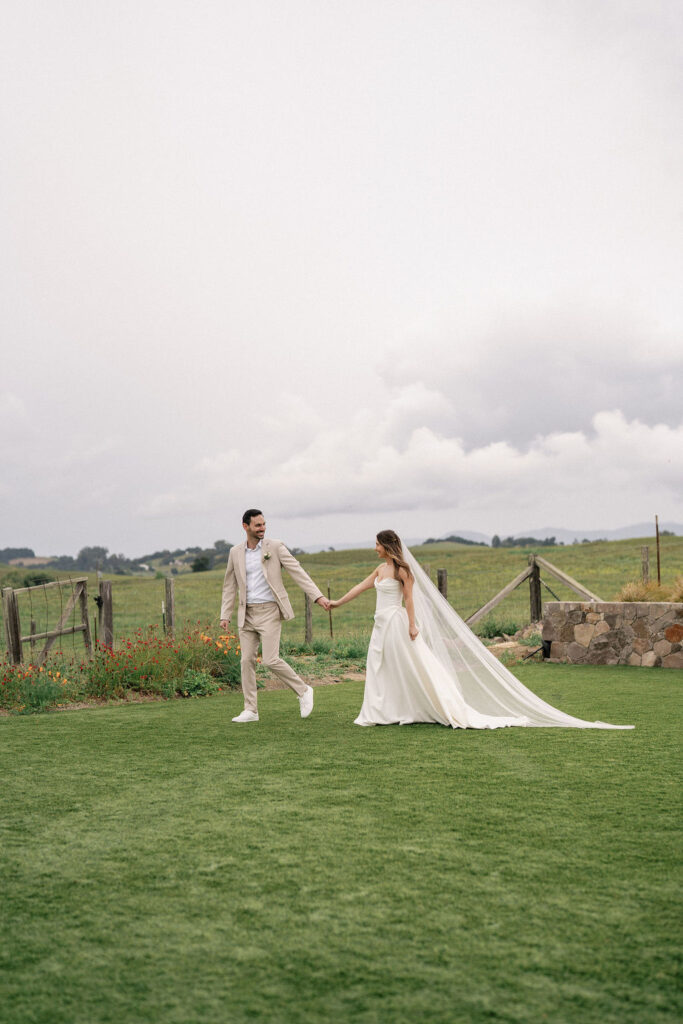 A bride and groom hold hands while walking on green grass outdoors, with a stone fence and cloudy sky in the background.