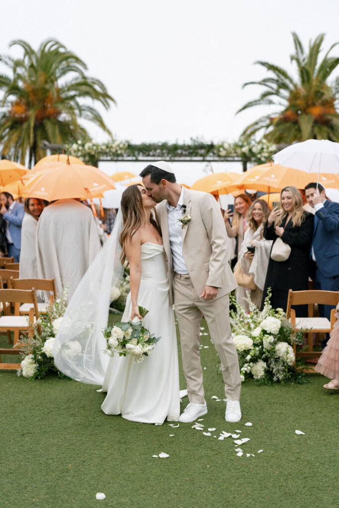 A bride and groom kiss at an outdoor wedding ceremony, surrounded by guests holding orange and white umbrellas, with palm trees in the background.
