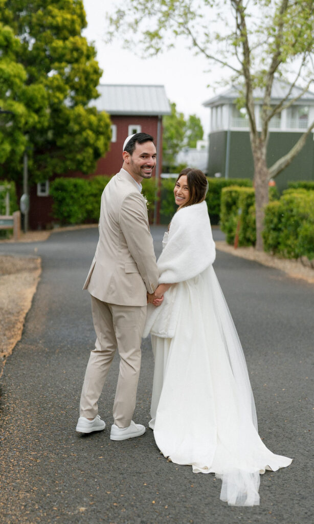 A bride and groom stand hand in hand on a paved path outdoors, both looking back and smiling at the camera. The bride wears a white dress and shawl; the groom is in a beige suit.