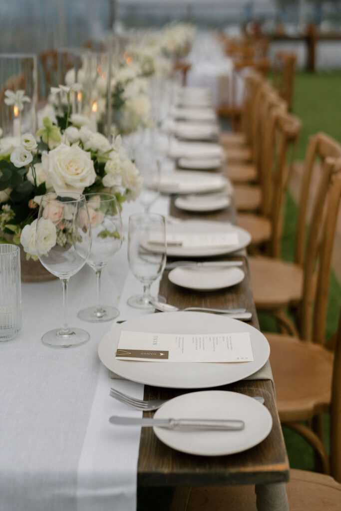 A long table set for an event with white plates, menus, glassware, silverware, and floral centerpieces featuring white roses on a white tablecloth.