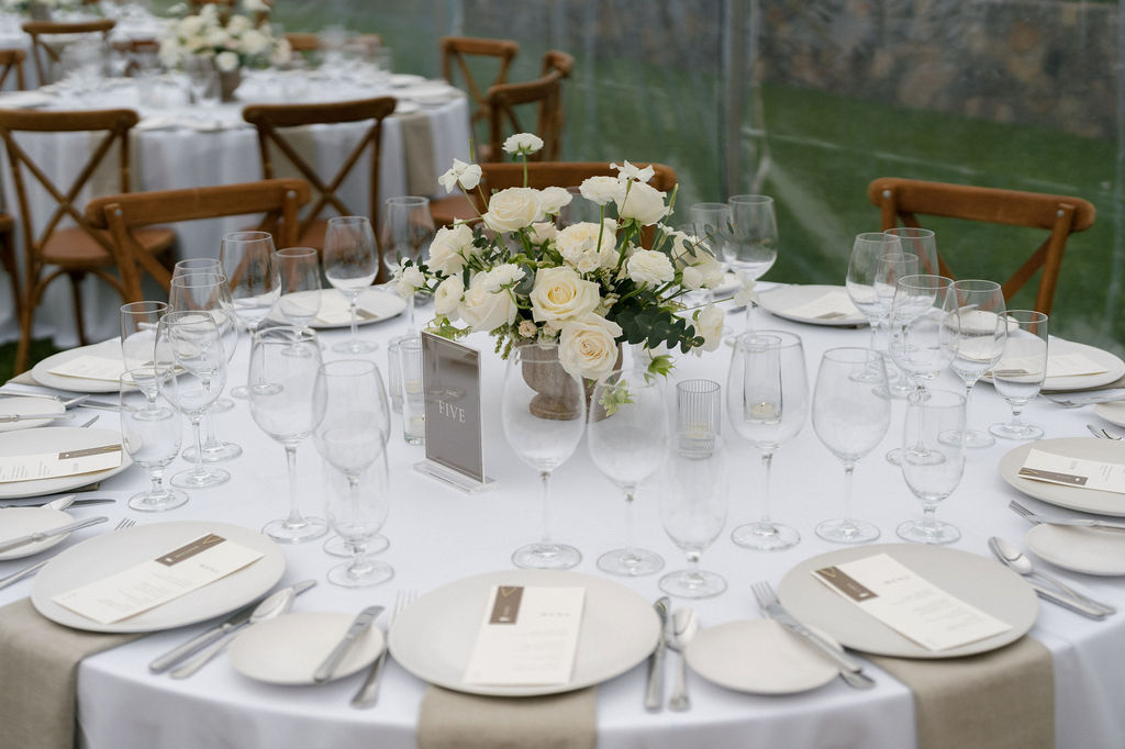 A round table set for an event with white tablecloth, glassware, plates, menus, beige napkins, and a centerpiece of white roses and greenery. Wooden chairs surround the table.