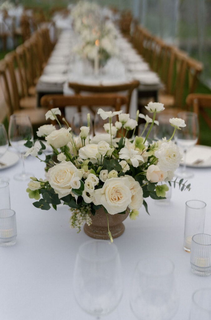 A white floral centerpiece with roses and greenery sits on a long, formally set dining table with wooden chairs and glassware.