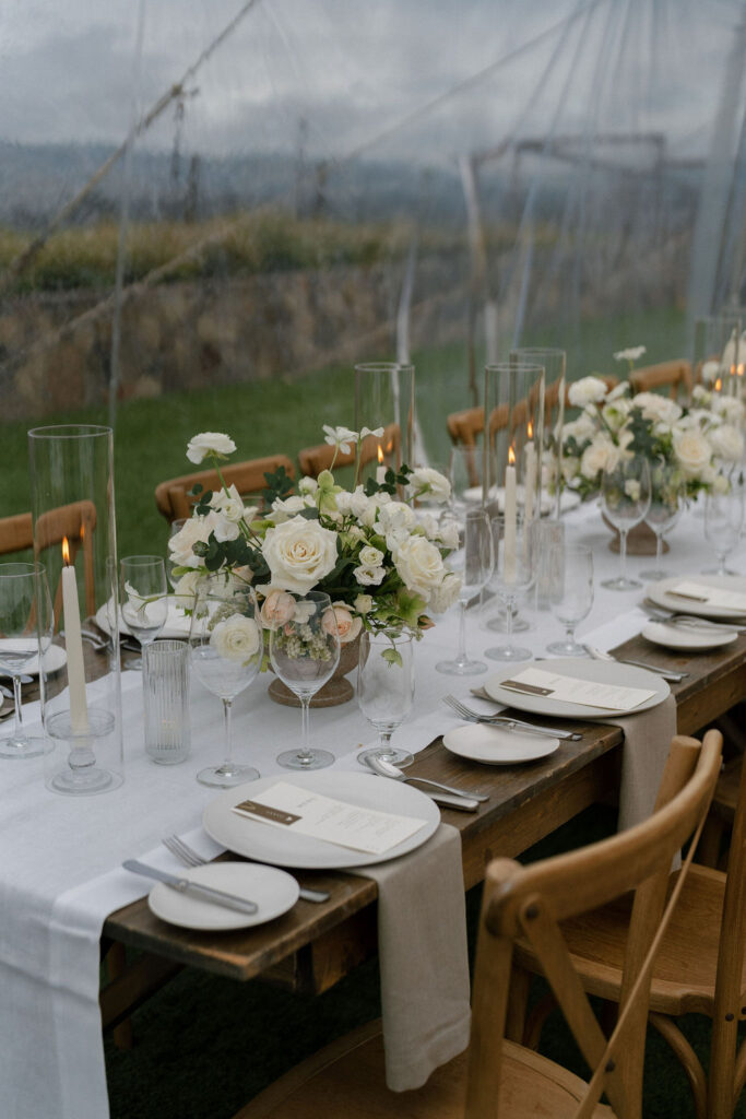 A long wooden table set for a formal event with white flowers, glass candle holders, menus, plates, and glasses on a white table runner, under a transparent tent.