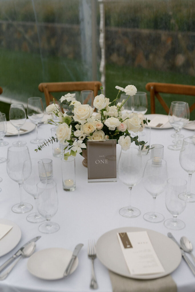 A round table set for an event with white flowers in a vase, a table number card, clear glassware, white plates, cutlery, and menus on a white tablecloth.
