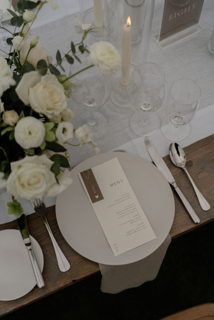 A neutral-toned table setting with white flowers, candles, glassware, cutlery, and a printed menu card placed on a round plate.