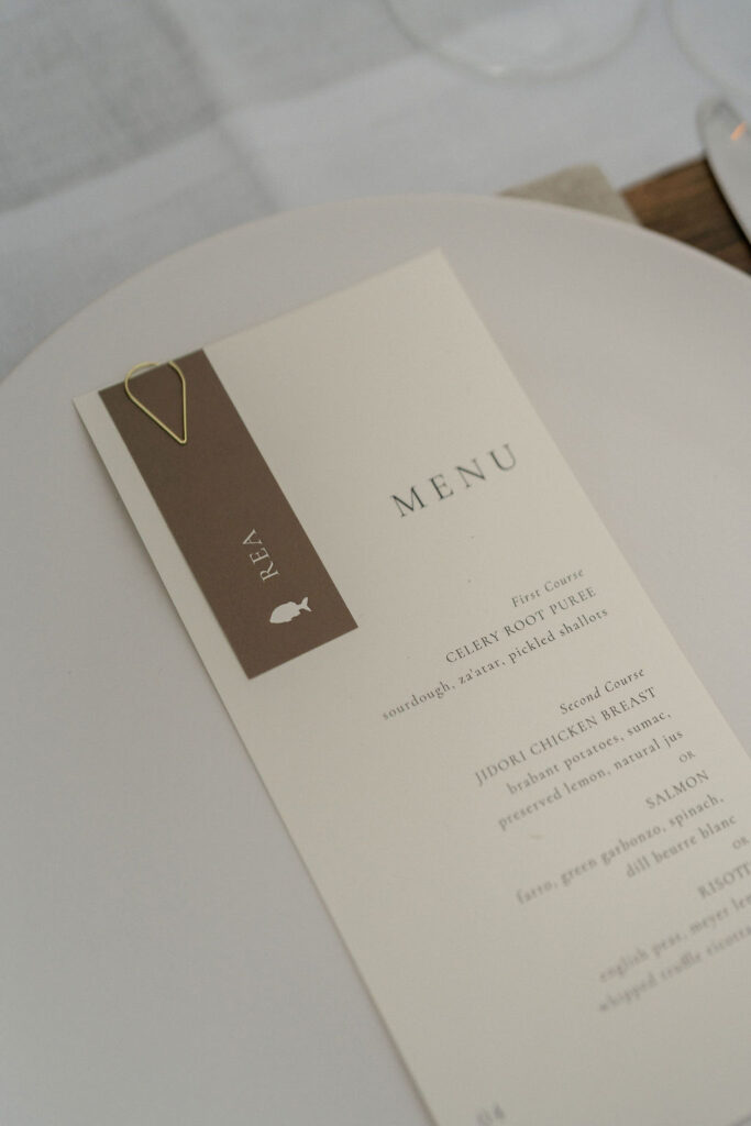 A beige and brown restaurant menu with a gold paperclip rests on a white plate, listing courses including chicken and salmon.