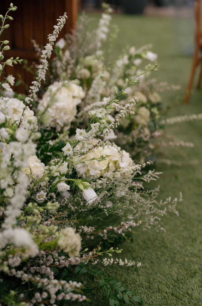 White flowers and greenery arranged closely together sit on a patch of green grass, with wooden furniture partially visible in the background.