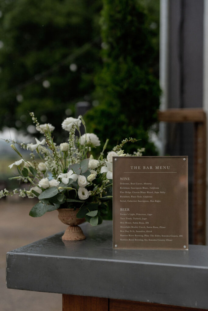 A floral arrangement in a brown vase sits next to a clear sign displaying a bar menu with wine and beer options on a metal countertop.