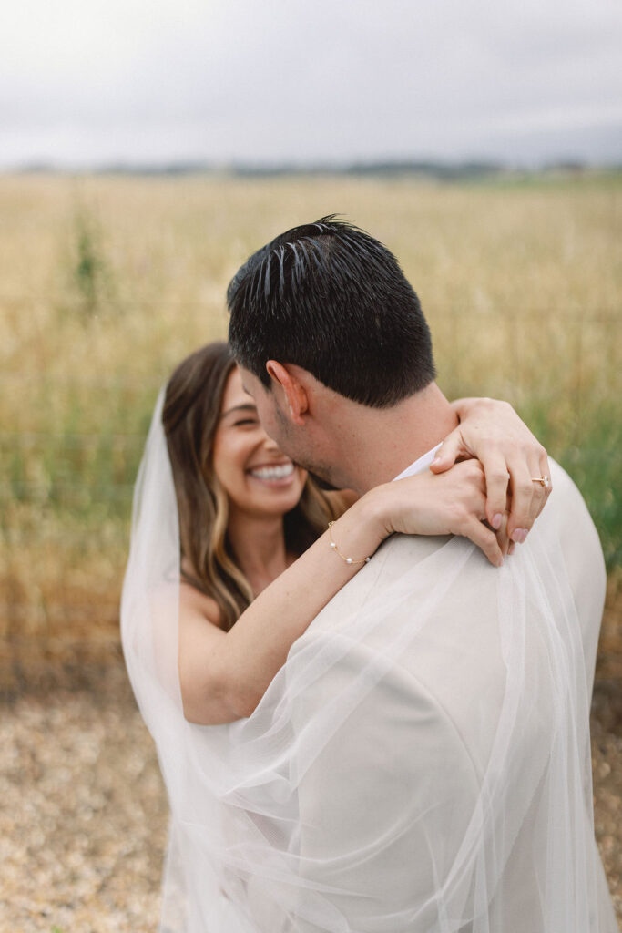 A bride and groom embrace outdoors, smiling, with the bride wearing a veil and wrapping her arms around the groom’s neck. A field stretches out in the background.